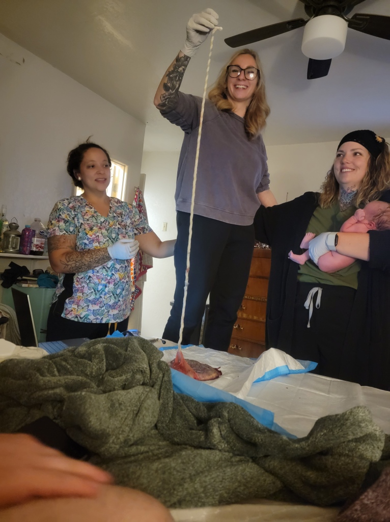 Three women stand at the foot of a bed. The one on the right holds a newborn baby. The one in the middle is standing on something to make her much taller than the other two, and holds up an umbilical cord to show it's length. It's about 4 feet long. 